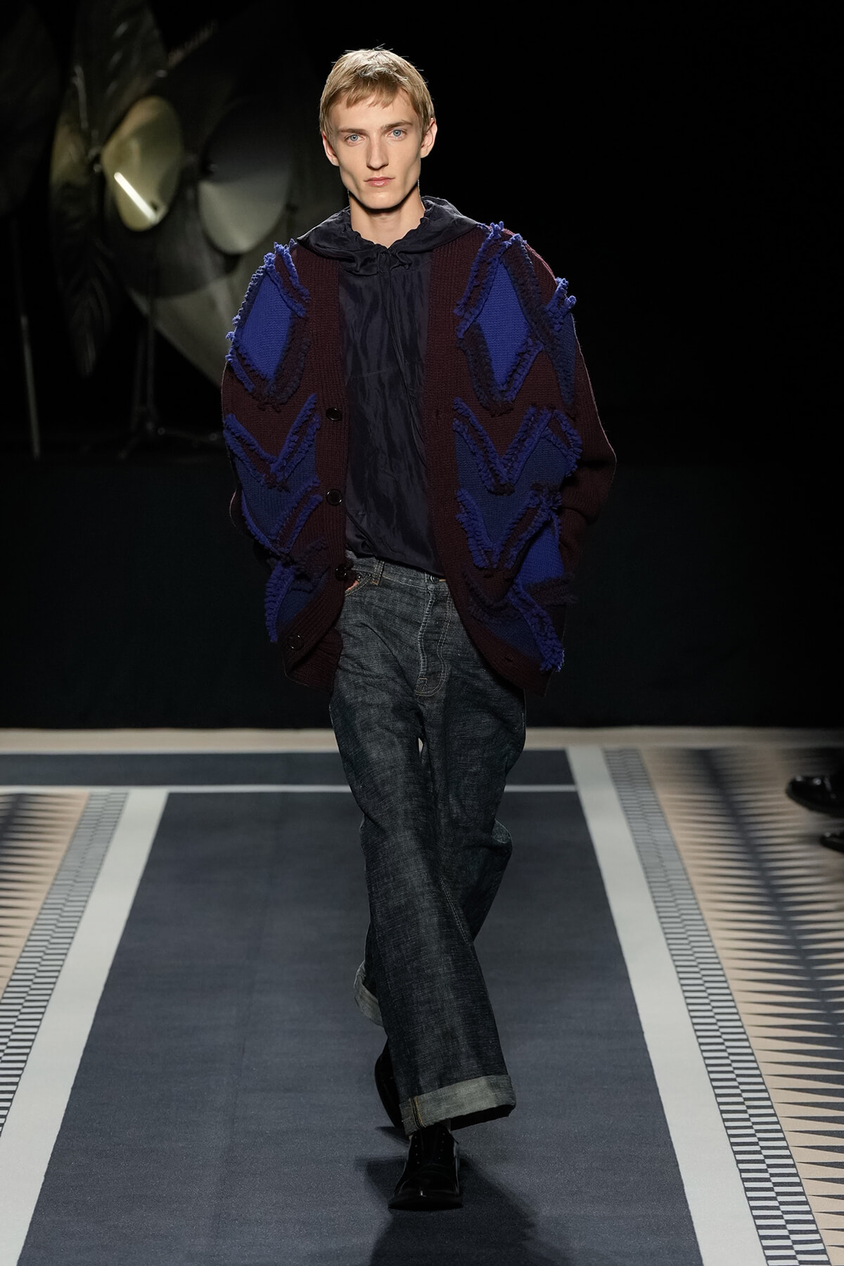 Male model walking runway in oversized maroon and blue cardigan with bold chevron knit, navy hoodie, wide dark denim jeans, black shoes