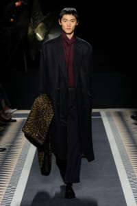 Male model struts a runway in a navy double-breasted coat over a burgundy shirt, carrying a leopard print coat.