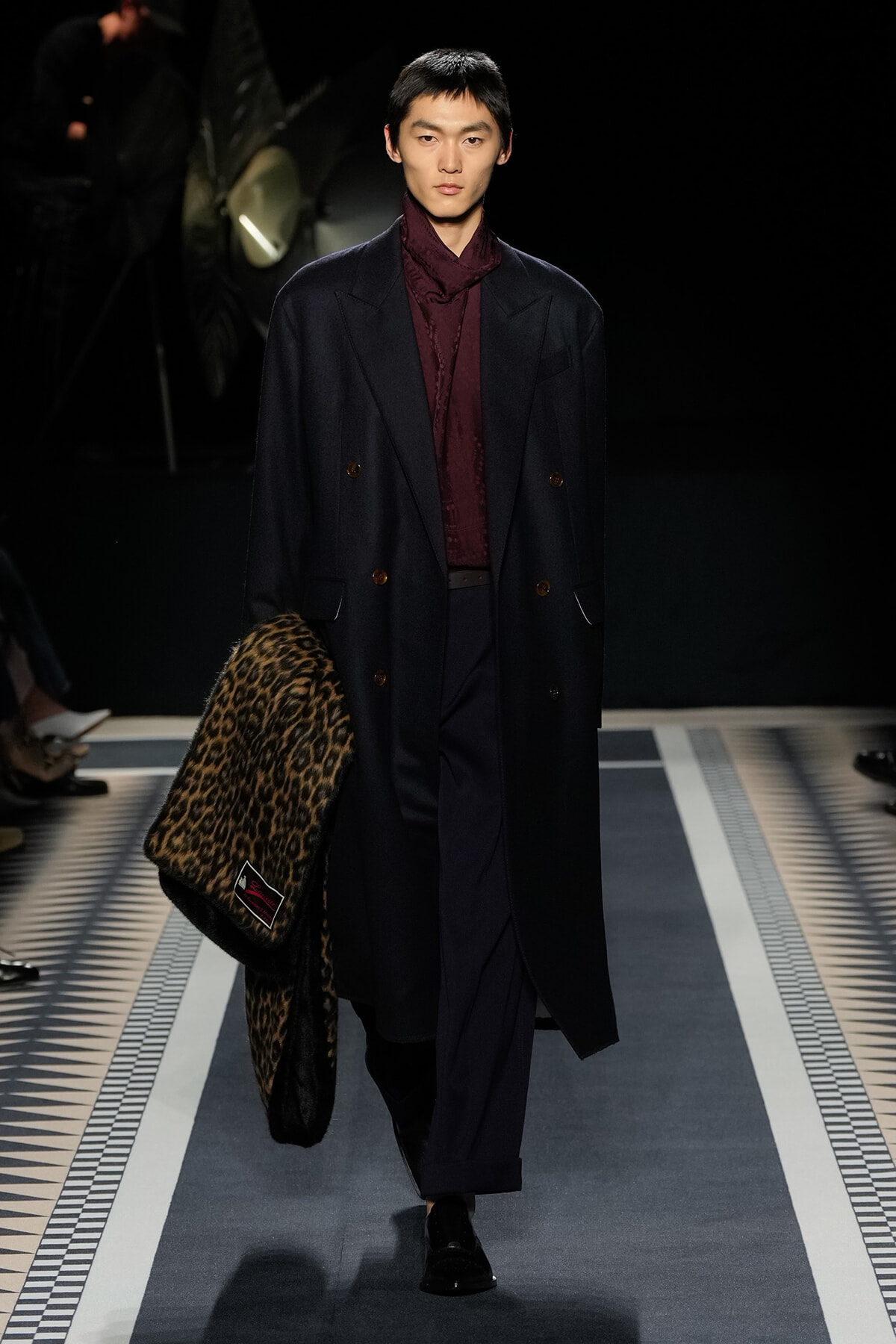 Male model struts a runway in a navy double-breasted coat over a burgundy shirt, carrying a leopard print coat.