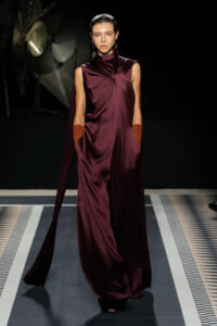 Model walks a fashion runway wearing a burgundy draped satin gown with a cape and long gloves, posing mid-stride against a dark backdrop.