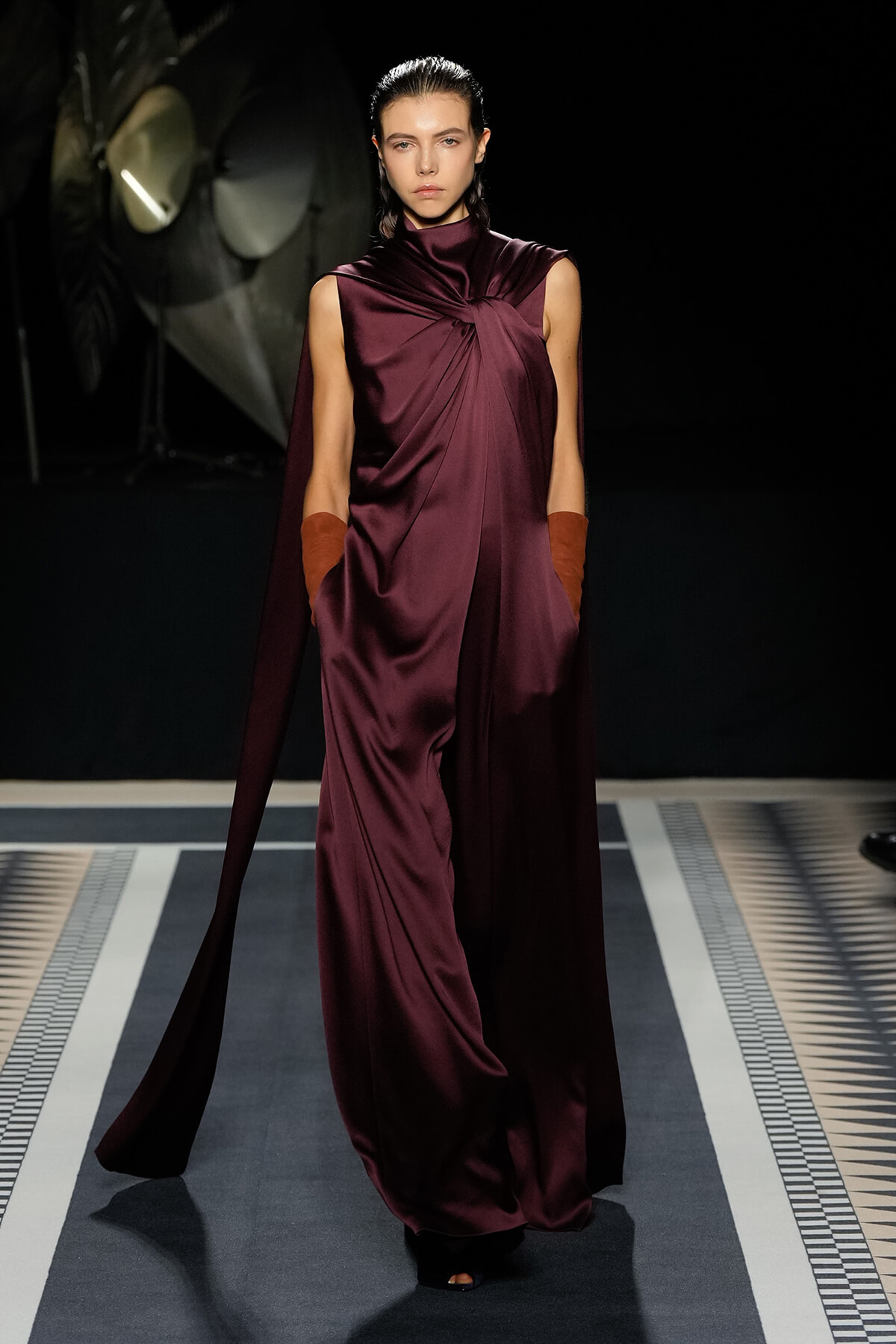 Model walks a fashion runway wearing a burgundy draped satin gown with a cape and long gloves, posing mid-stride against a dark backdrop.