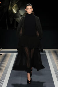 Model walking a runway in an all-black ensemble with a high-neck top, sheer puff sleeves, and a voluminous layered skirt, black heels visible.