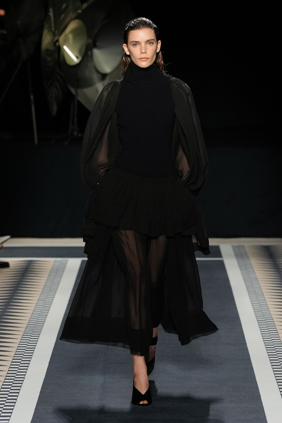 Model walking a runway in an all-black ensemble with a high-neck top, sheer puff sleeves, and a voluminous layered skirt, black heels visible.