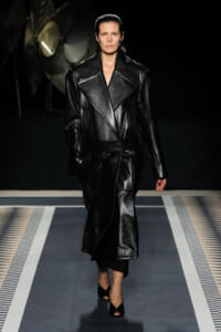 Model walking a runway in a glossy black leather trench with wide lapels and belt, dark background.