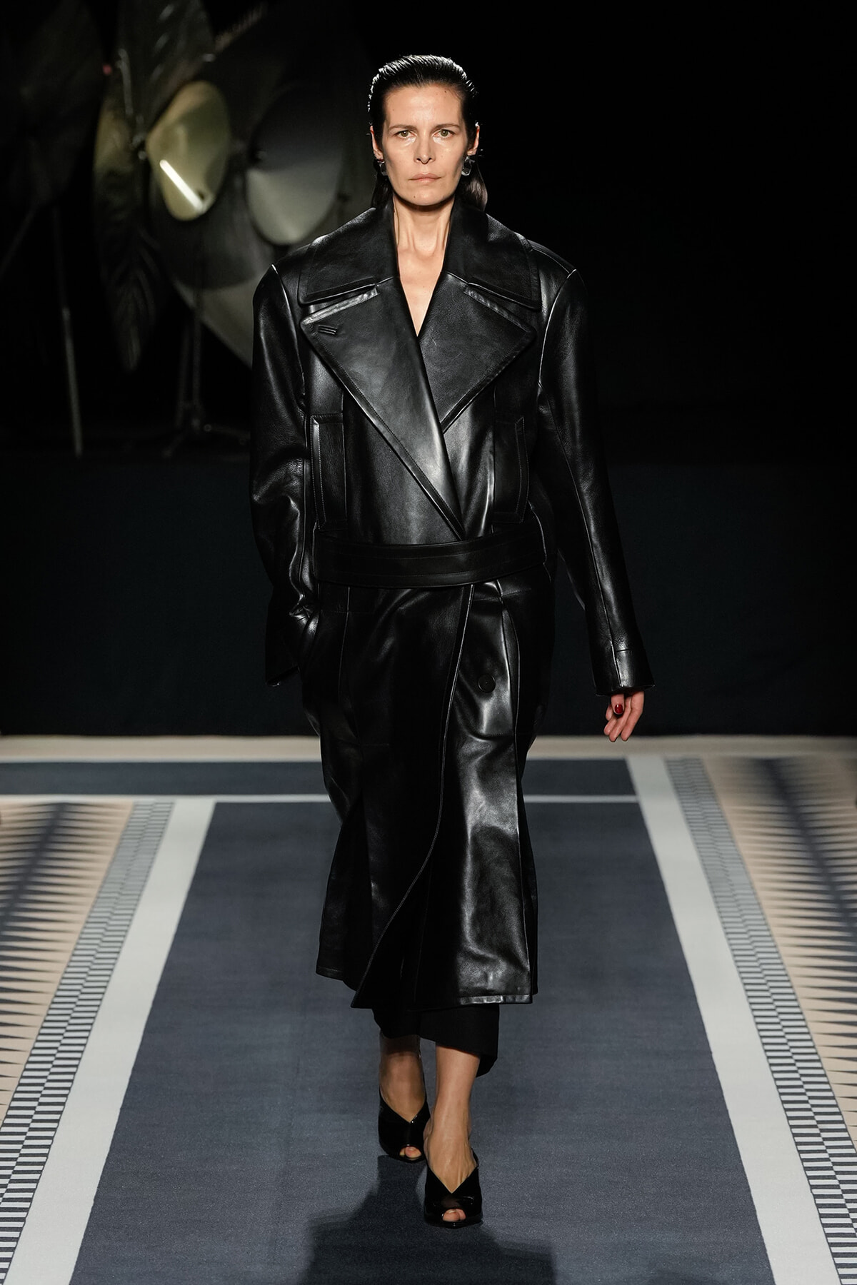 Model walking a runway in a glossy black leather trench with wide lapels and belt, dark background.