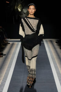 Model walks a runway in a monochrome geometric ensemble: sheer knit top with black panels, cream sleeves, a black belt, and patterned wide-leg pants with matching boots.