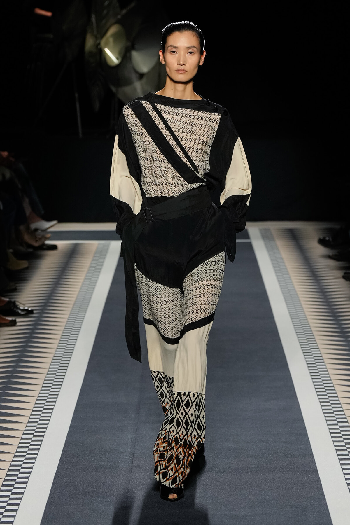 Model walks a runway in a monochrome geometric ensemble: sheer knit top with black panels, cream sleeves, a black belt, and patterned wide-leg pants with matching boots.