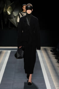 Model walks a fashion runway in an all-black textured coat, turtleneck, blindfold-style headband, and carries a black geometric bag.