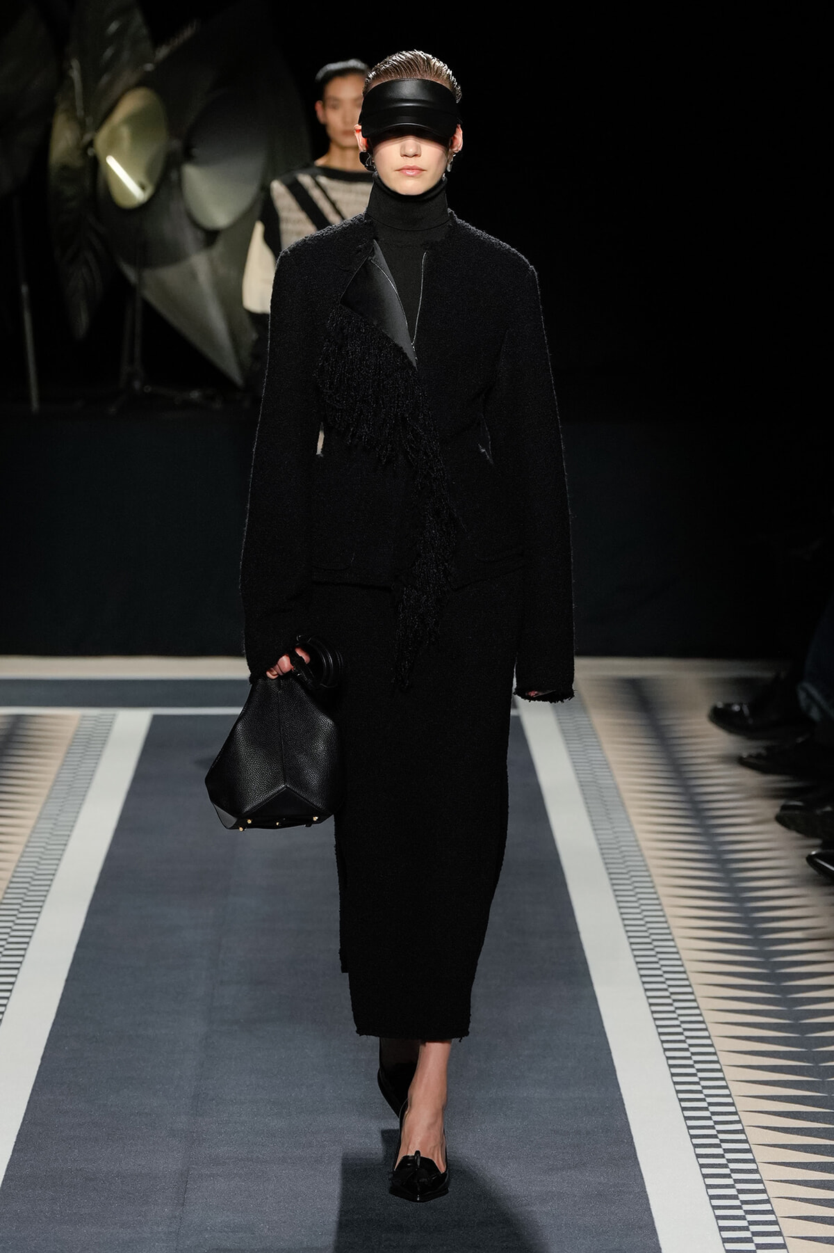 Model walks a fashion runway in an all-black textured coat, turtleneck, blindfold-style headband, and carries a black geometric bag.