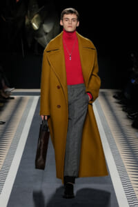 Model on a runway wearing a camel oversized coat over a red turtleneck and gray plaid trousers, carrying a dark tote bag. Necklace pendant visible.