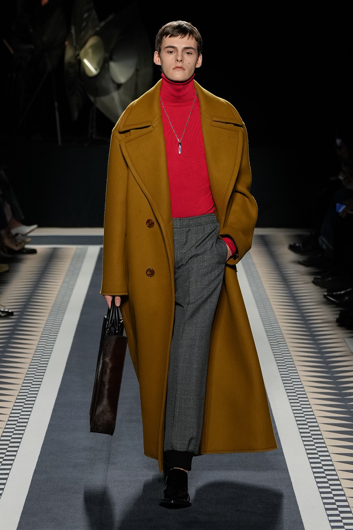 Model on a runway wearing a camel oversized coat over a red turtleneck and gray plaid trousers, carrying a dark tote bag. Necklace pendant visible.