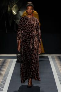 Model walking a runway in a brown zigzag-print, long-sleeve maxidress with a waist belt, paired with black heels and a small black handbag.