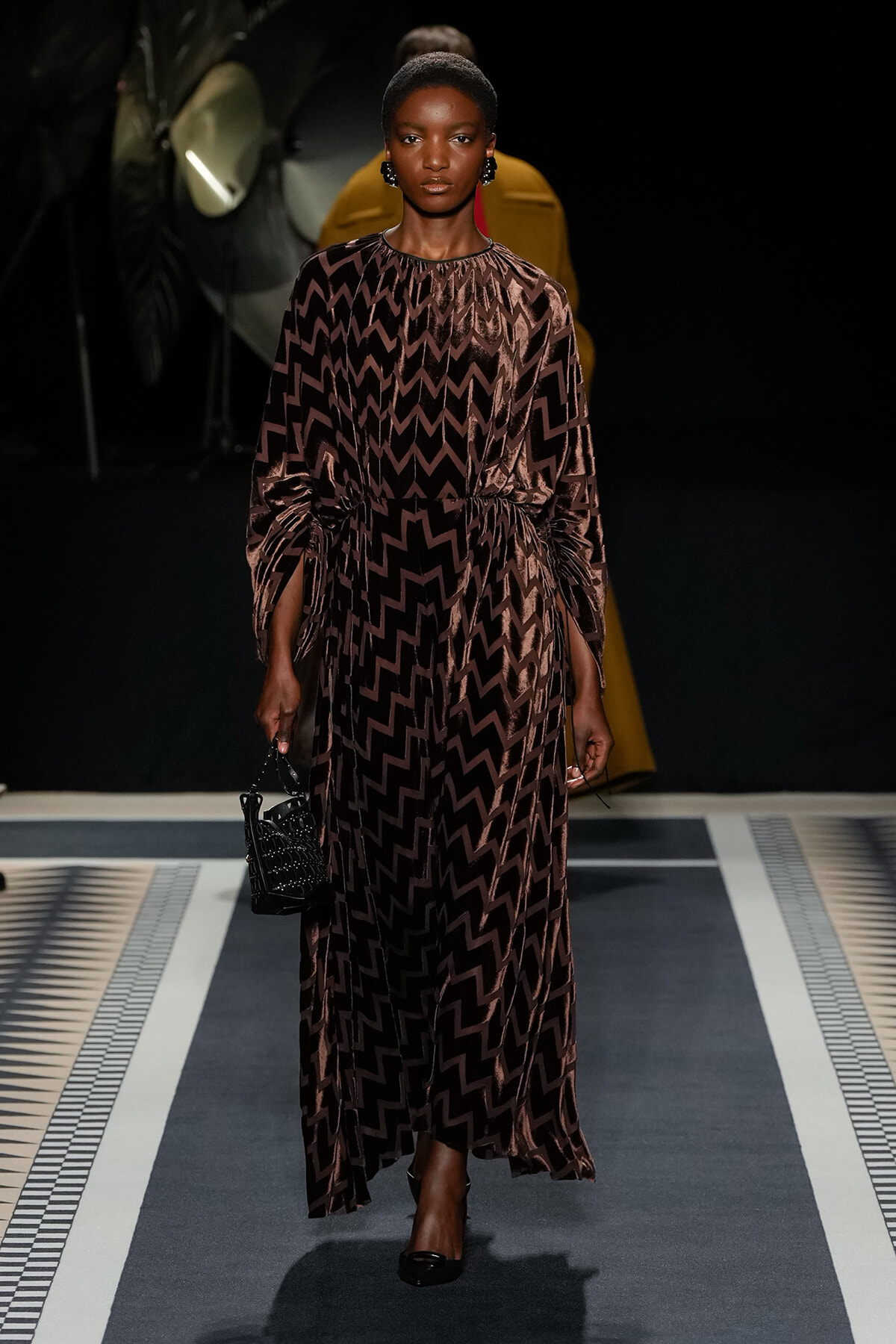 Model walking a runway in a brown zigzag-print, long-sleeve maxidress with a waist belt, paired with black heels and a small black handbag.