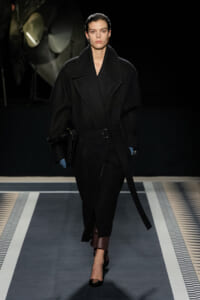 Model walking a runway in an oversized black wool coat with wide lapels and a belt, carrying a black bag and wearing black heels against a dark backdrop.