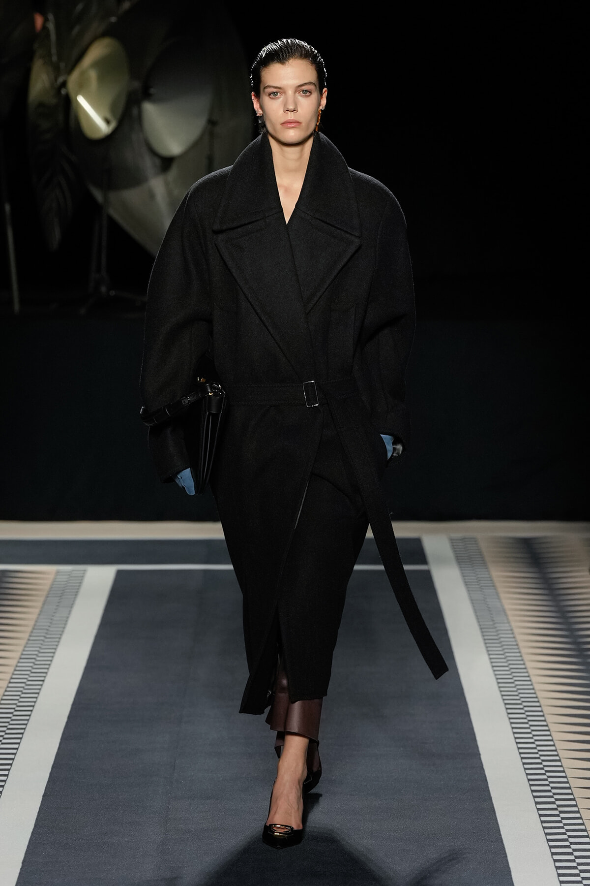 Model walking a runway in an oversized black wool coat with wide lapels and a belt, carrying a black bag and wearing black heels against a dark backdrop.