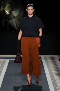 Model on a runway wearing a navy blouse with puff sleeves and burnt orange wide-legged trousers, carrying a burgundy handbag.