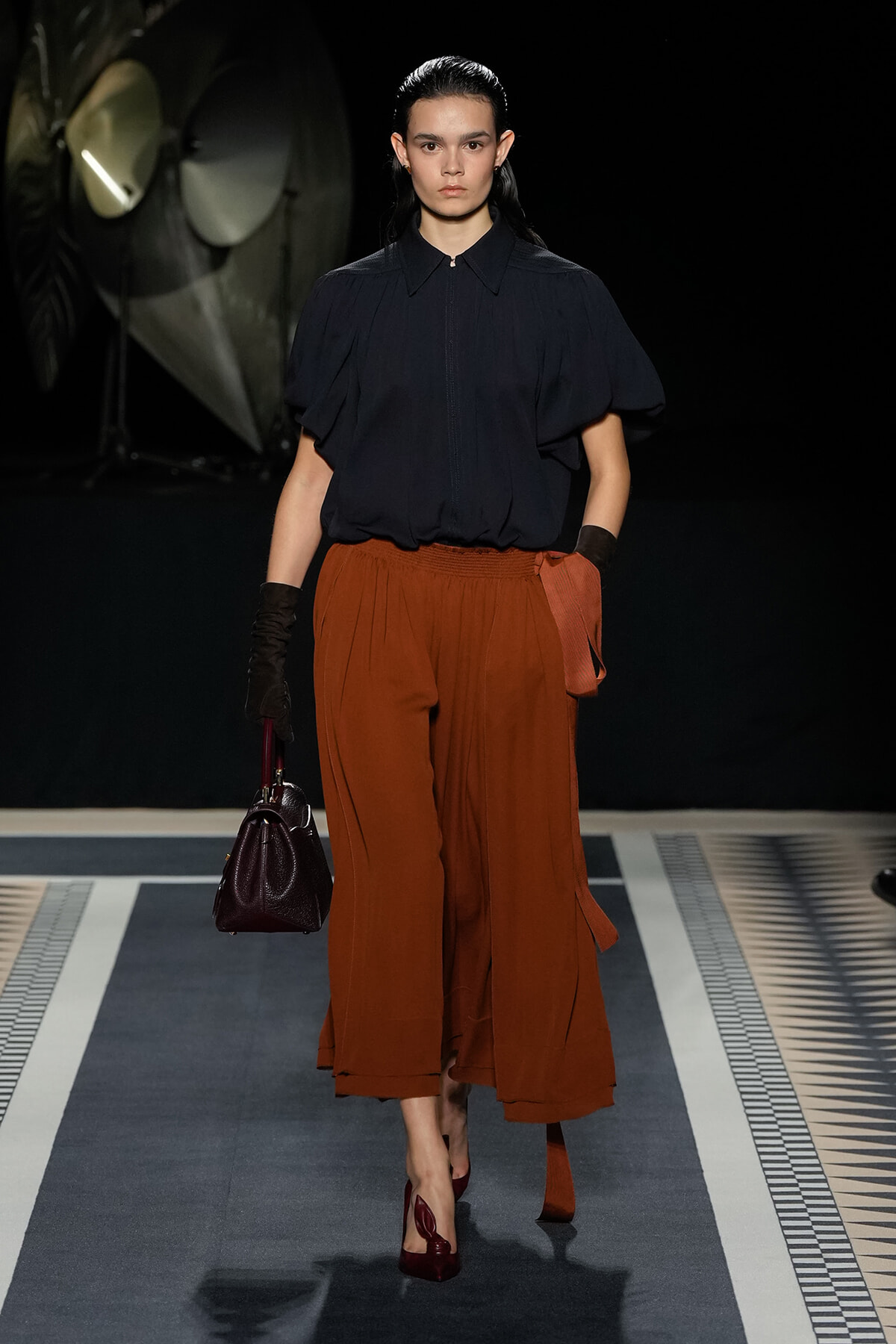 Model on a runway wearing a navy blouse with puff sleeves and burnt orange wide-legged trousers, carrying a burgundy handbag.