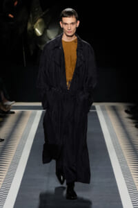 Male model walking the runway in a long black trench over a mustard blouse, hands in pockets, dark backdrop.