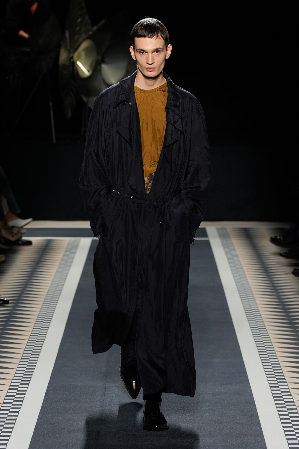 Male model walking the runway in a long black trench over a mustard blouse, hands in pockets, dark backdrop.