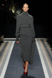 Model walks a fashion runway in a monochrome gray knit turtleneck and matching wrap skirt, with burgundy open-toe heels