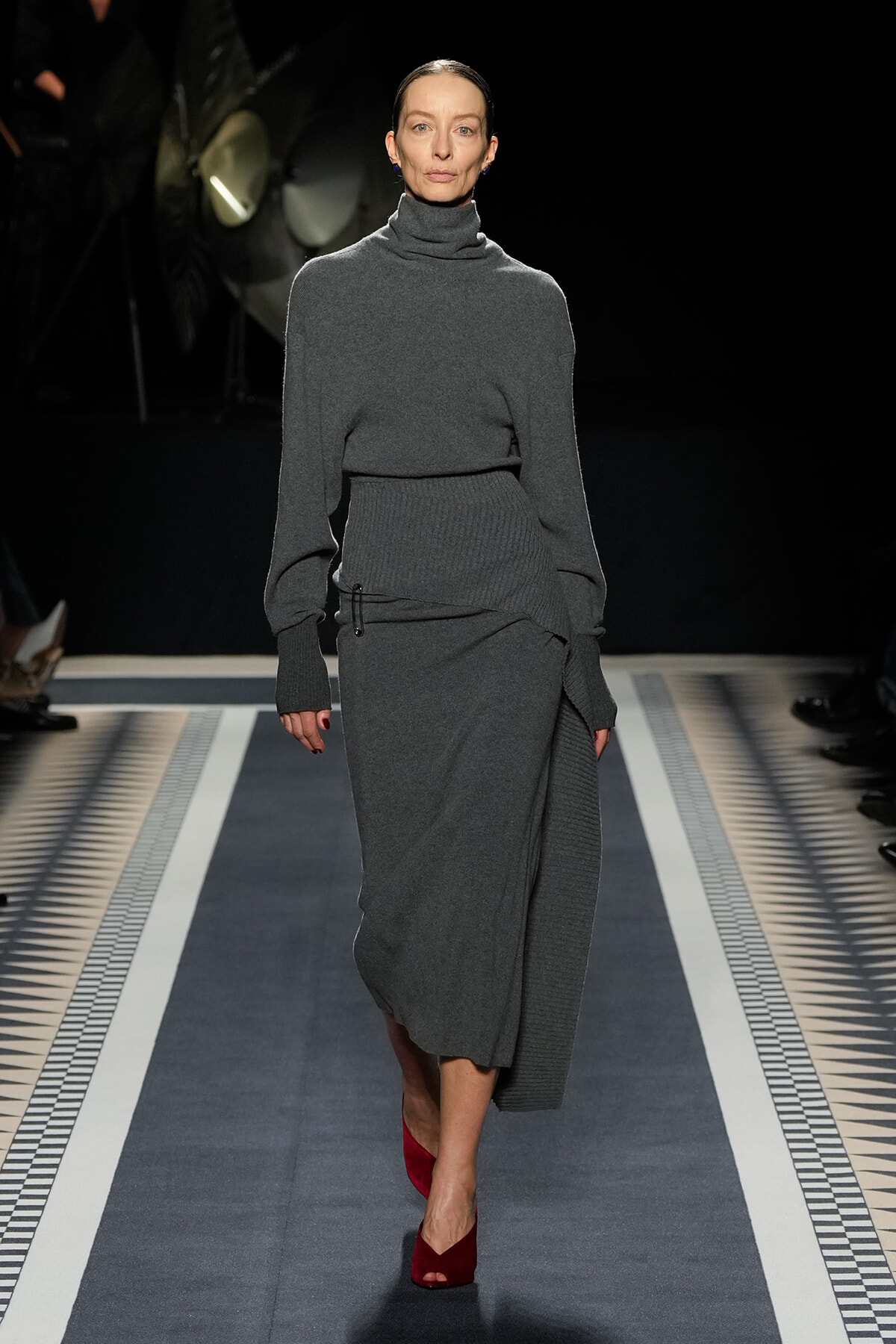 Model walks a fashion runway in a monochrome gray knit turtleneck and matching wrap skirt, with burgundy open-toe heels