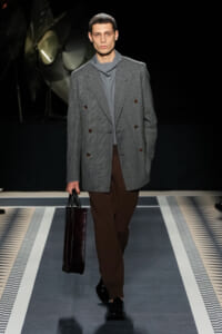 Male model walks a runway in a gray plaid double-breasted blazer, gray turtleneck, and brown trousers, carrying a dark rectangular briefcase.