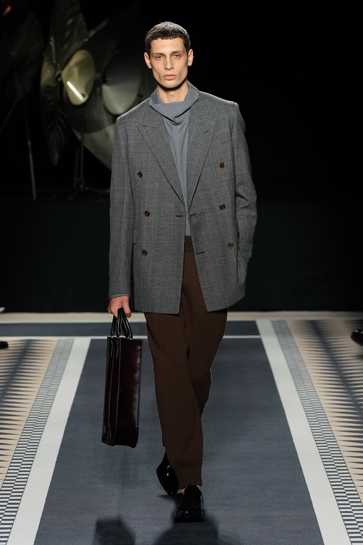 Male model walks a runway in a gray plaid double-breasted blazer, gray turtleneck, and brown trousers, carrying a dark rectangular briefcase.