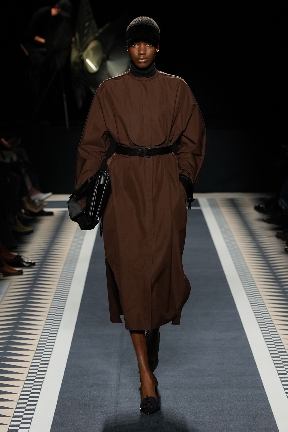 Model on a runway wearing a brown belted trench dress, black belt, gloves, headband, and clutch bag.