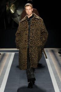 Model walks the runway in a long leopard-print faux fur coat over dark denim and black shirt, spotlighted on a fashion catwalk.