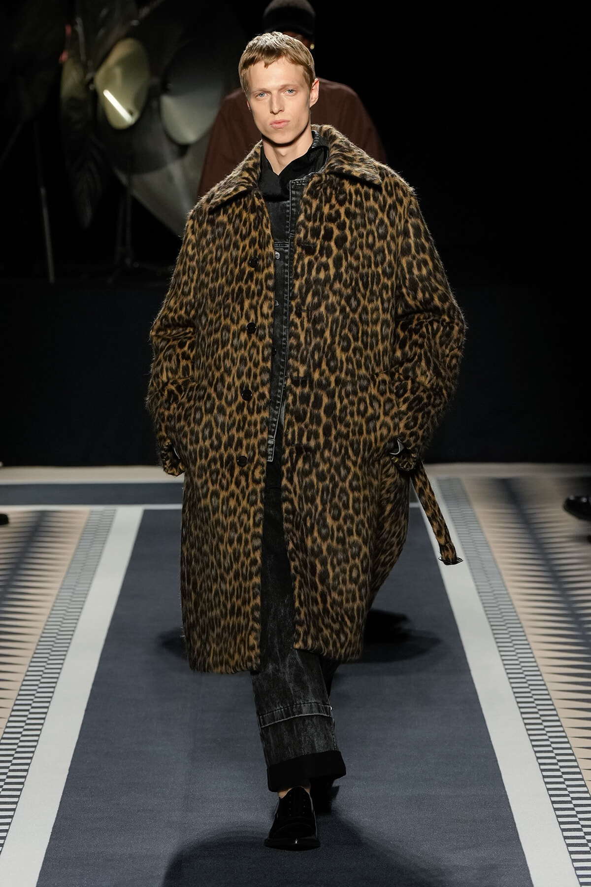 Model walks the runway in a long leopard-print faux fur coat over dark denim and black shirt, spotlighted on a fashion catwalk.