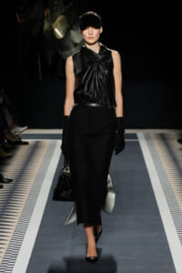 Model walks a black monochrome runway wearing a draped black sleeveless top, black pencil skirt, gloves, cap, and a black handbag