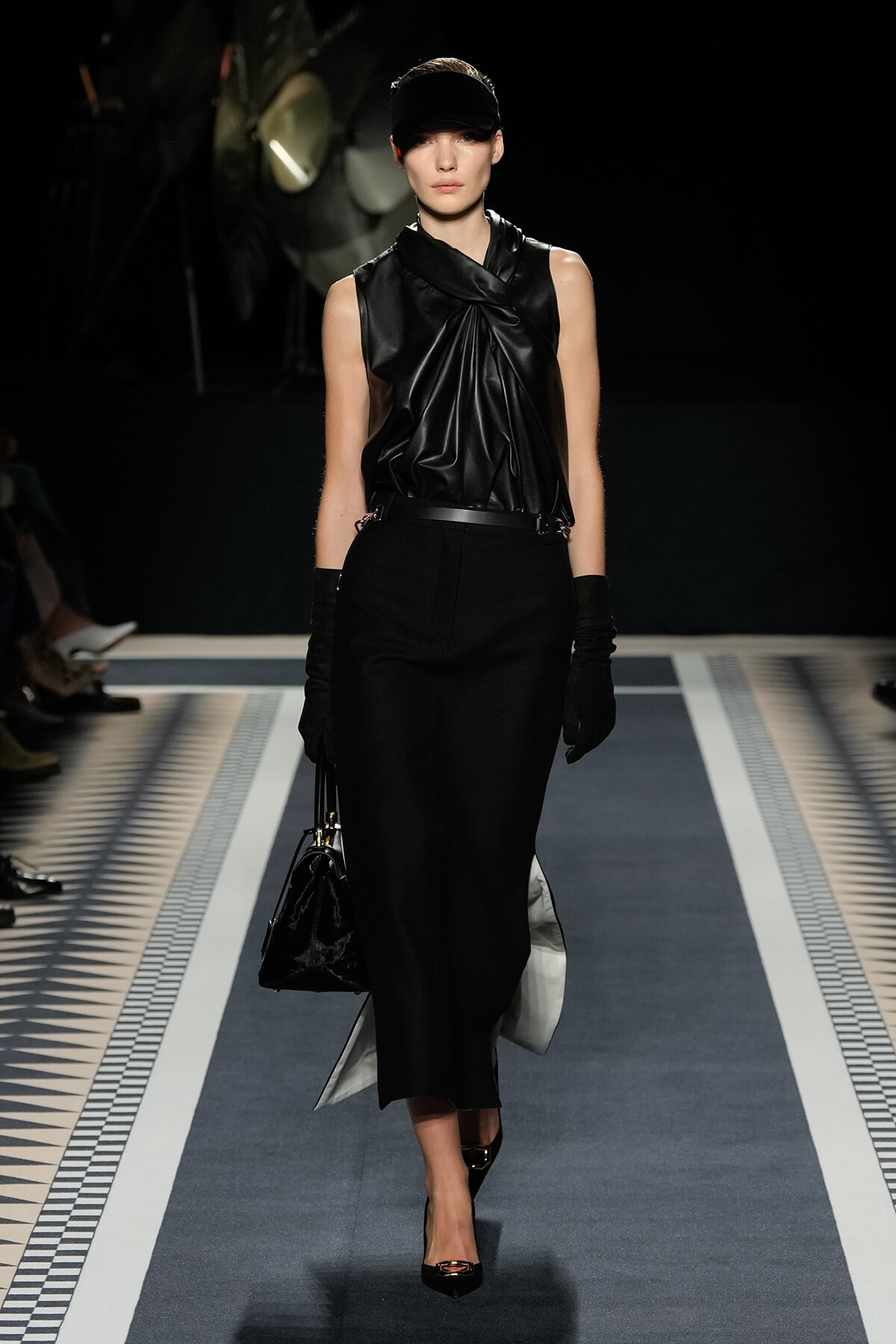 Model walks a black monochrome runway wearing a draped black sleeveless top, black pencil skirt, gloves, cap, and a black handbag