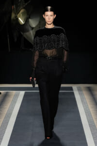 Runway model in black ensemble: sheer-sleeve top with a beaded cape over high-waisted trousers, carrying a small clutch.