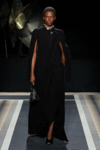 Model on a runway wearing a floor-length black textured cape dress with a slit, accessorized with a chunky necklace and a small black quilted handbag.
