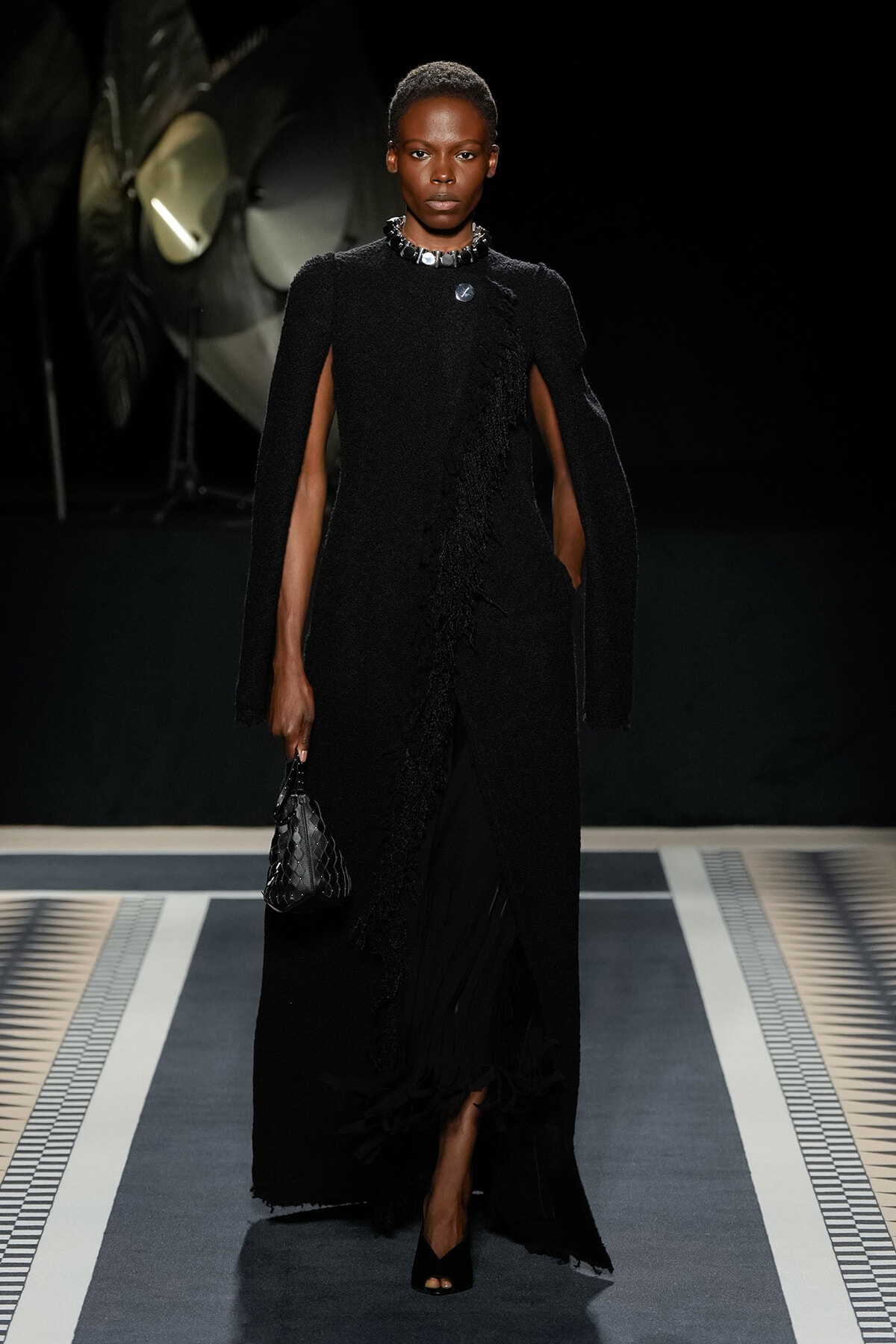 Model on a runway wearing a floor-length black textured cape dress with a slit, accessorized with a chunky necklace and a small black quilted handbag.
