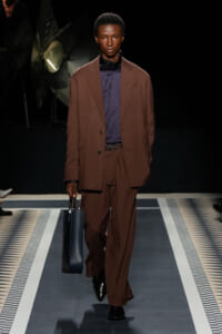 Runway model in a brown suit with a navy top, black belt, and a dark blue briefcase-style bag.