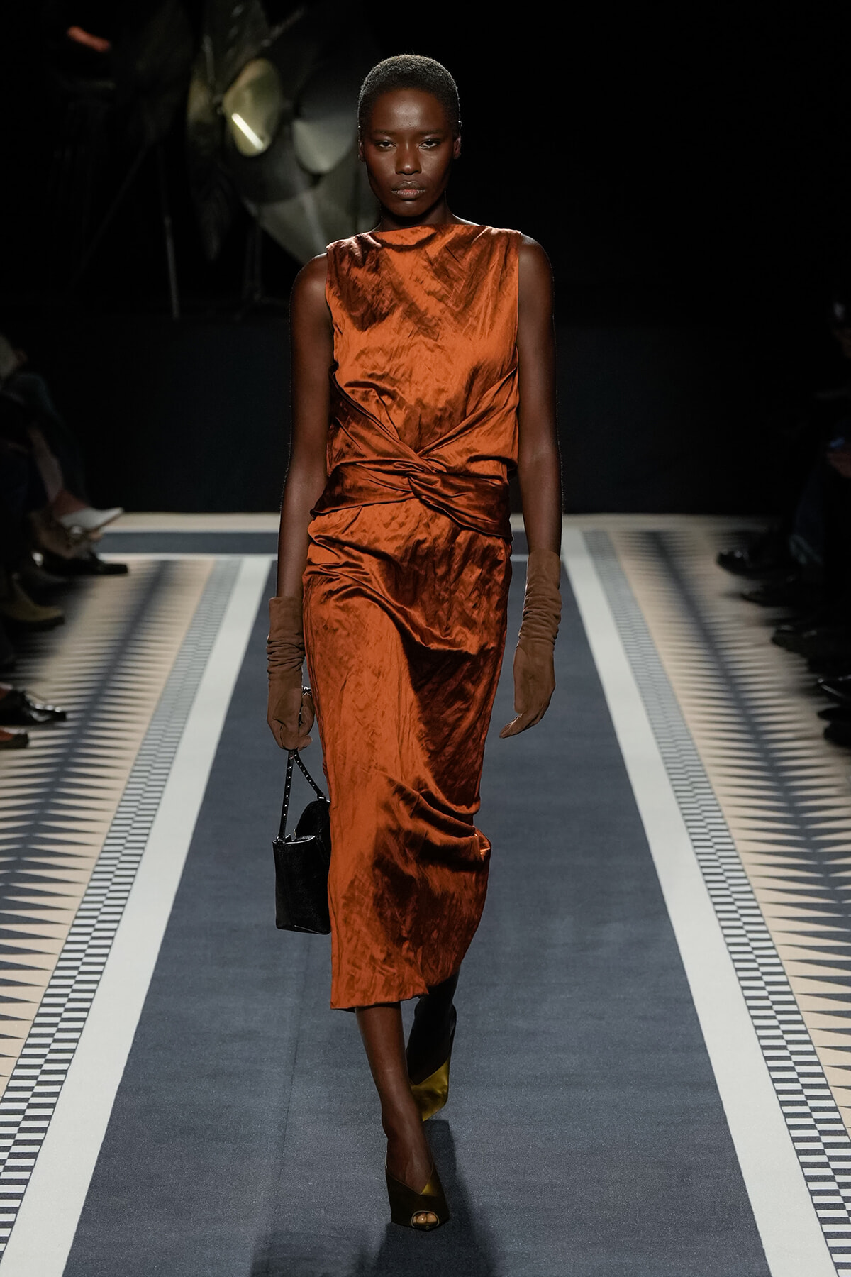 Model walks a runway in a rust-orange satin sleeveless dress with a twisted waist, brown gloves, and a small black handbag.
