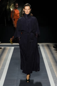 Model walks a fashion runway wearing a long navy trench coat with a belt, high collar and voluminous sleeves, gloves at the wrists.