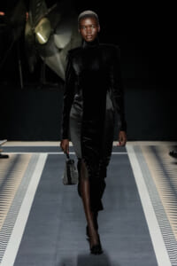 Model walks the runway in a black velvet turtleneck dress with structured shoulders, carrying a small patterned handbag and wearing black heels.
