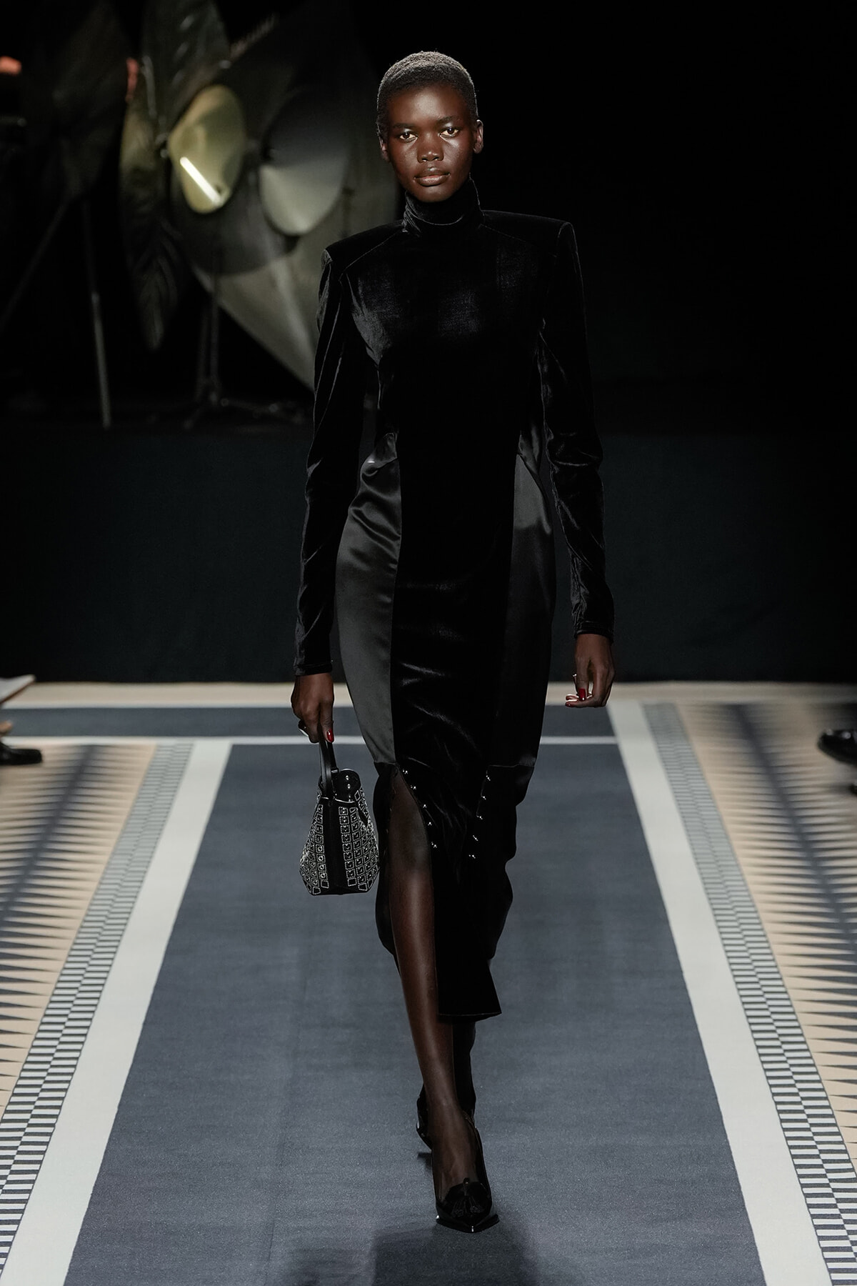 Model walks the runway in a black velvet turtleneck dress with structured shoulders, carrying a small patterned handbag and wearing black heels.