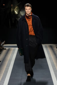 Male model walking on runway in a long navy coat over an orange turtleneck and dark trousers, holding a green clutch.