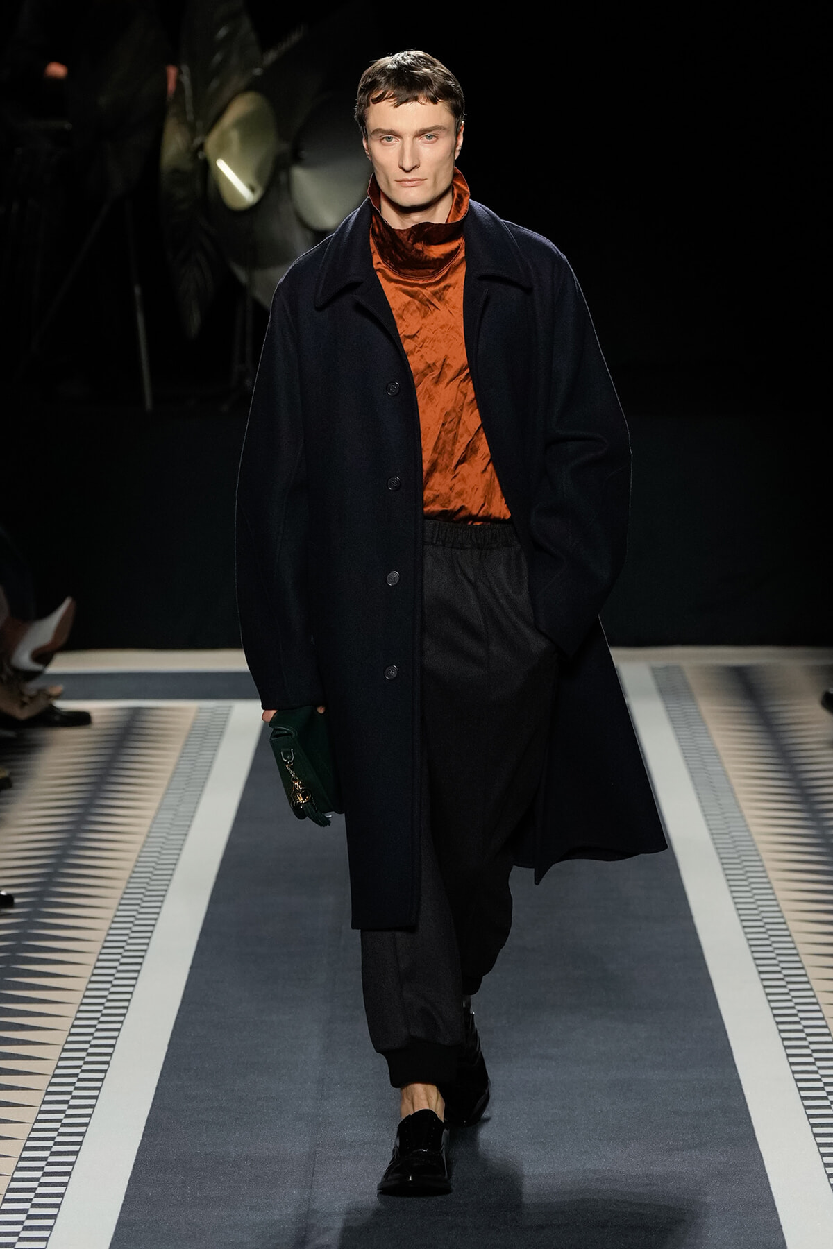Male model walking on runway in a long navy coat over an orange turtleneck and dark trousers, holding a green clutch.