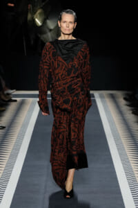 Model walking a fashion runway in a red-black patterned, draped dress with black accents and open-toe black heels, slicked-back hair.