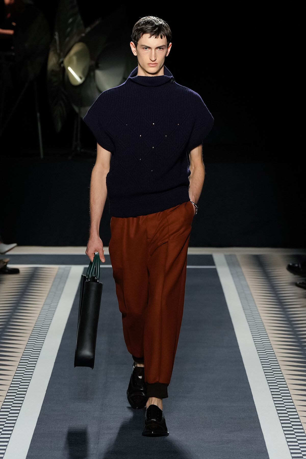 Male model walks a runway in a navy knit poncho-style turtleneck and rust brown wide-leg trousers, carrying a black tote with green handles.
