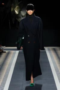 Model on a runway wearing a navy double-breasted coat, black turtleneck, dark visor hat, emerald green handbag, and matching green heels.