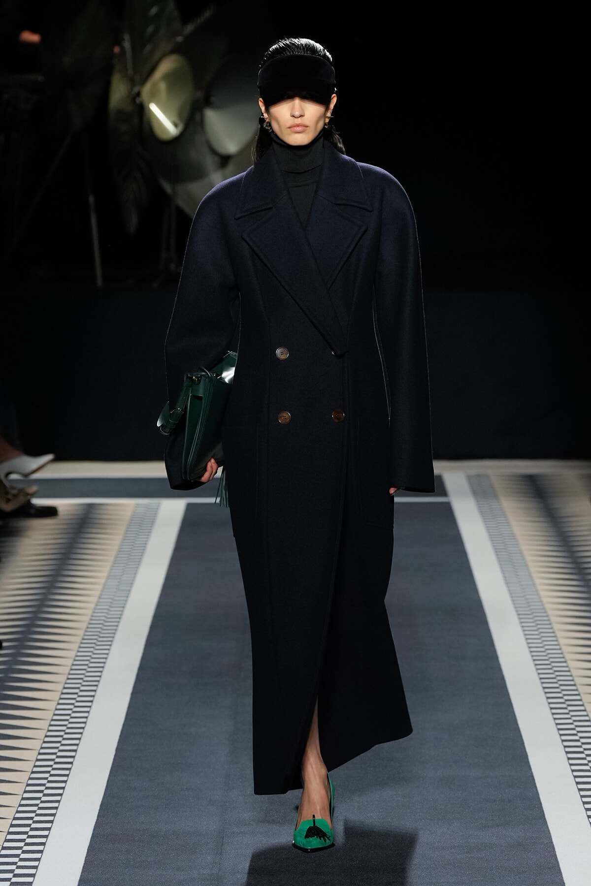 Model on a runway wearing a navy double-breasted coat, black turtleneck, dark visor hat, emerald green handbag, and matching green heels.