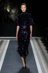 Model walking a runway in a navy, sequined midi dress with puff sleeves and high neck.