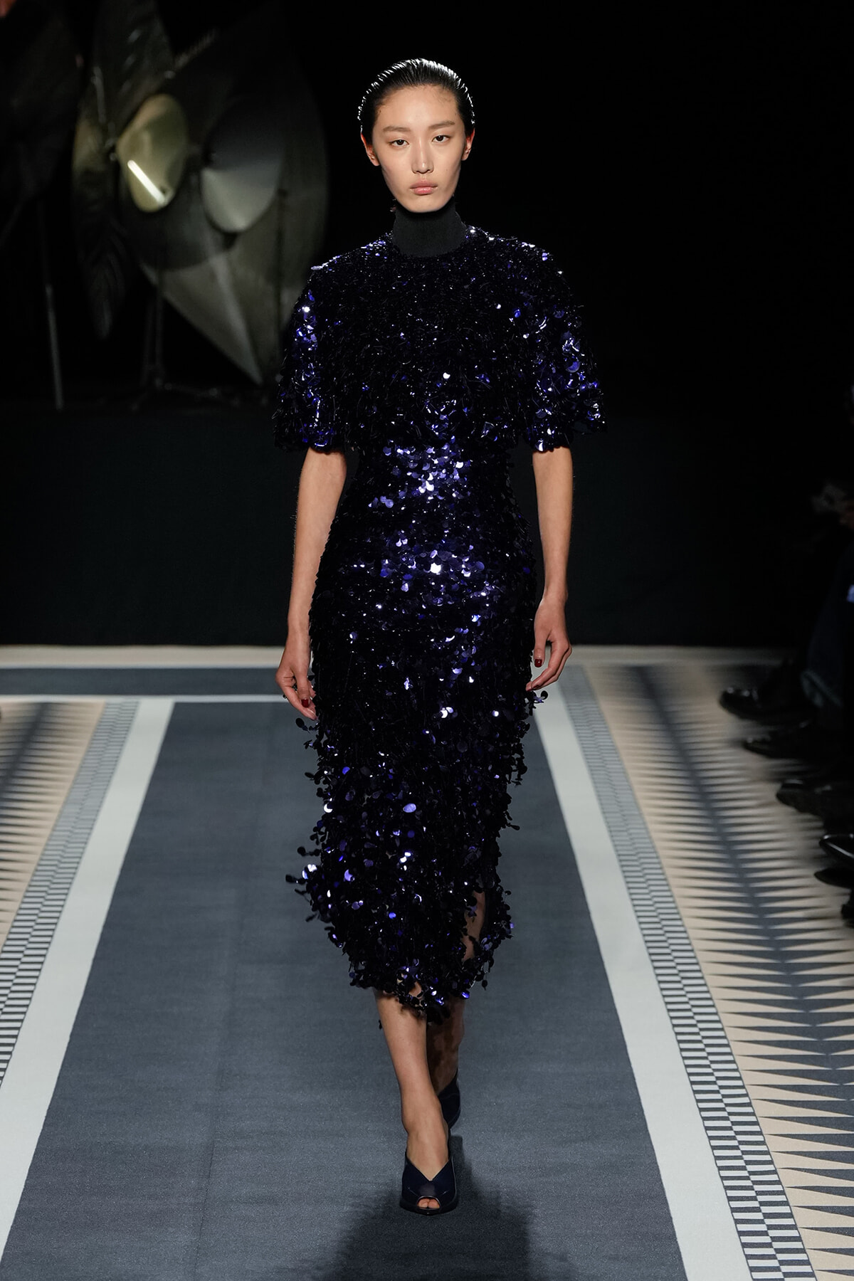 Model walking a runway in a navy, sequined midi dress with puff sleeves and high neck.