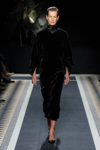 Model walking a fashion runway in a long black velvet dress with a high neck and gathered sleeves, black heels, under stage lighting.