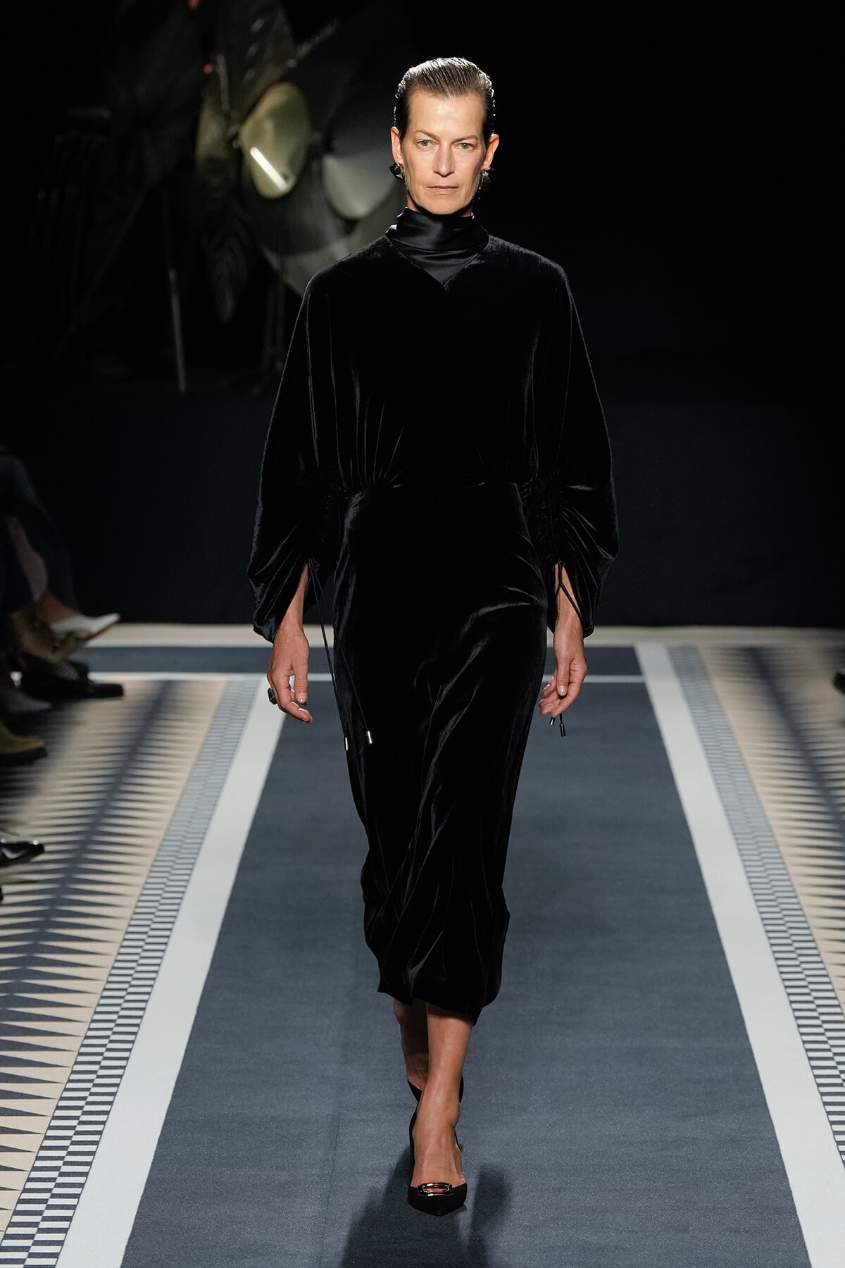 Model walking a fashion runway in a long black velvet dress with a high neck and gathered sleeves, black heels, under stage lighting.